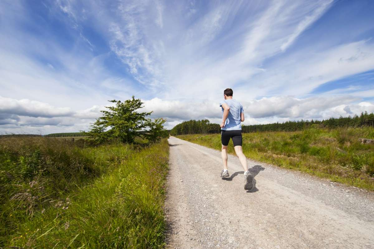 Man in light blue tee and black running shorts jogging along a gravel road under big sky